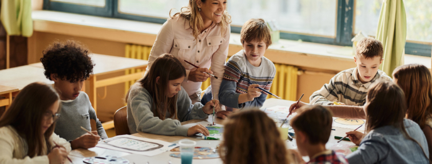 Eine Gruppe von Schüler:innen sitzt um einen großen Tisch und malt. Eine Lehrerin steht mit Pinsel in der Hand und lächelt einer Schülerin zu.