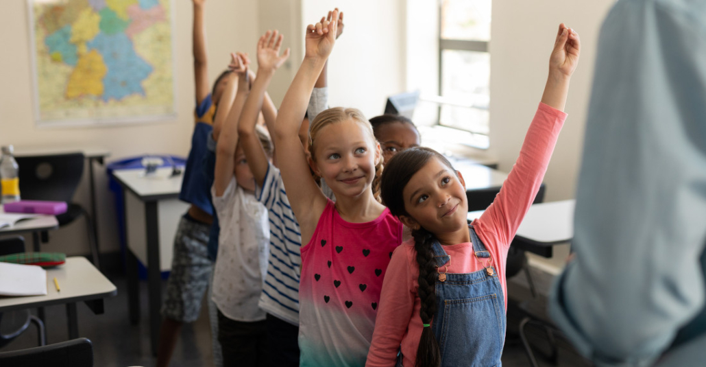 Volksschulkinder stehen in einem Klassenzimmer in einer Reihe hintereinander und heben die Hände in die Höhe