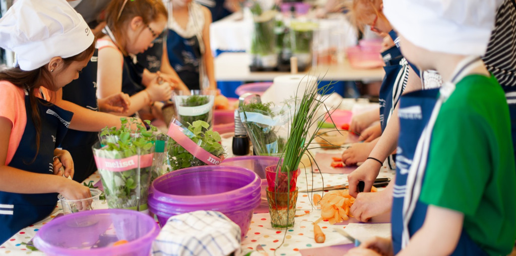 Kinder mit Kochmützen stehen an einem Tisch und schneiden Gemüse