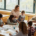 Eine Gruppe von Schüler:innen sitzt um einen großen Tisch und malt. Eine Lehrerin steht mit Pinsel in der Hand und lächelt einer Schülerin zu.